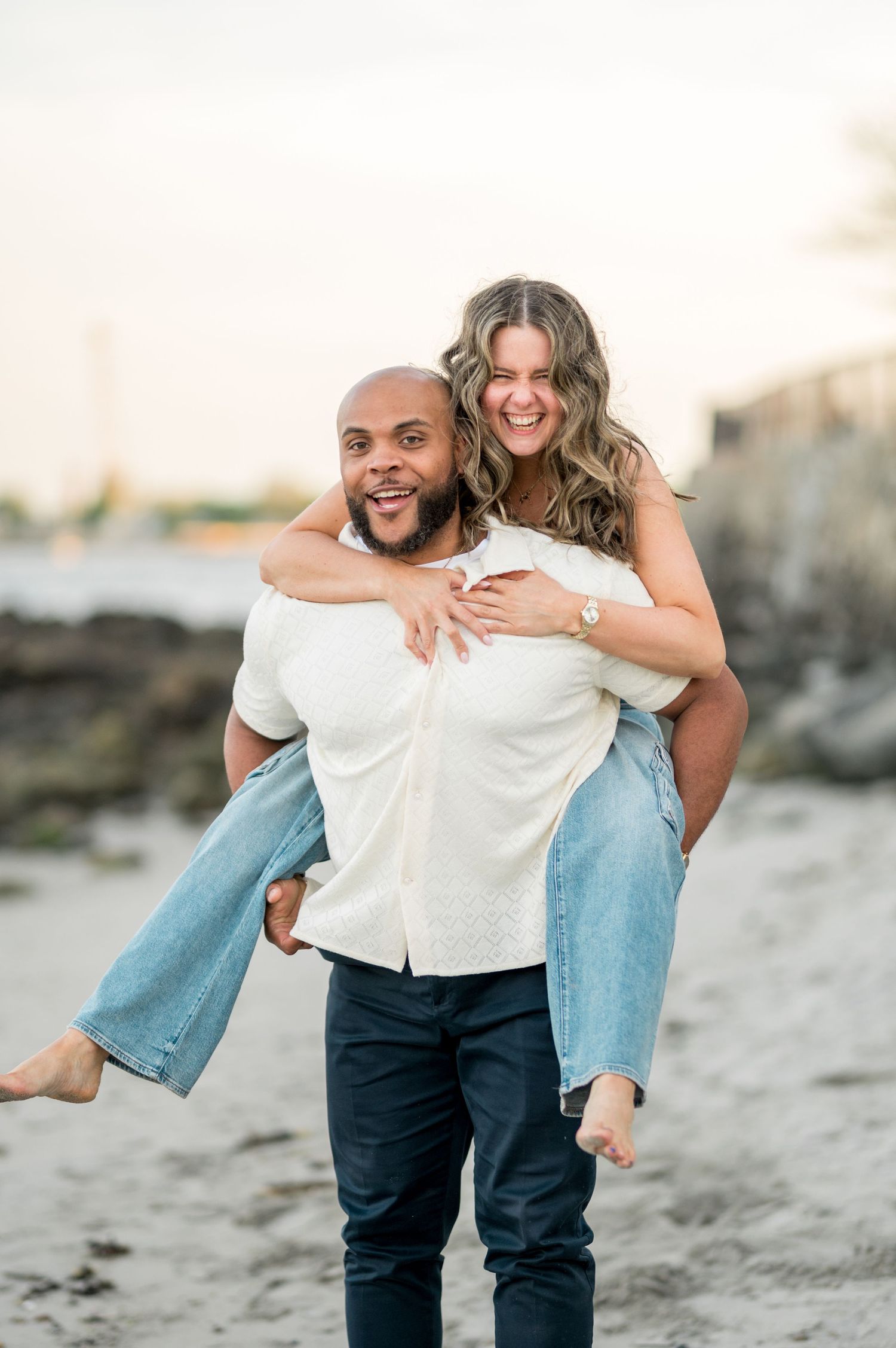 A playful couple embraces in a piggyback pose during a beach photoshoot at sunset.