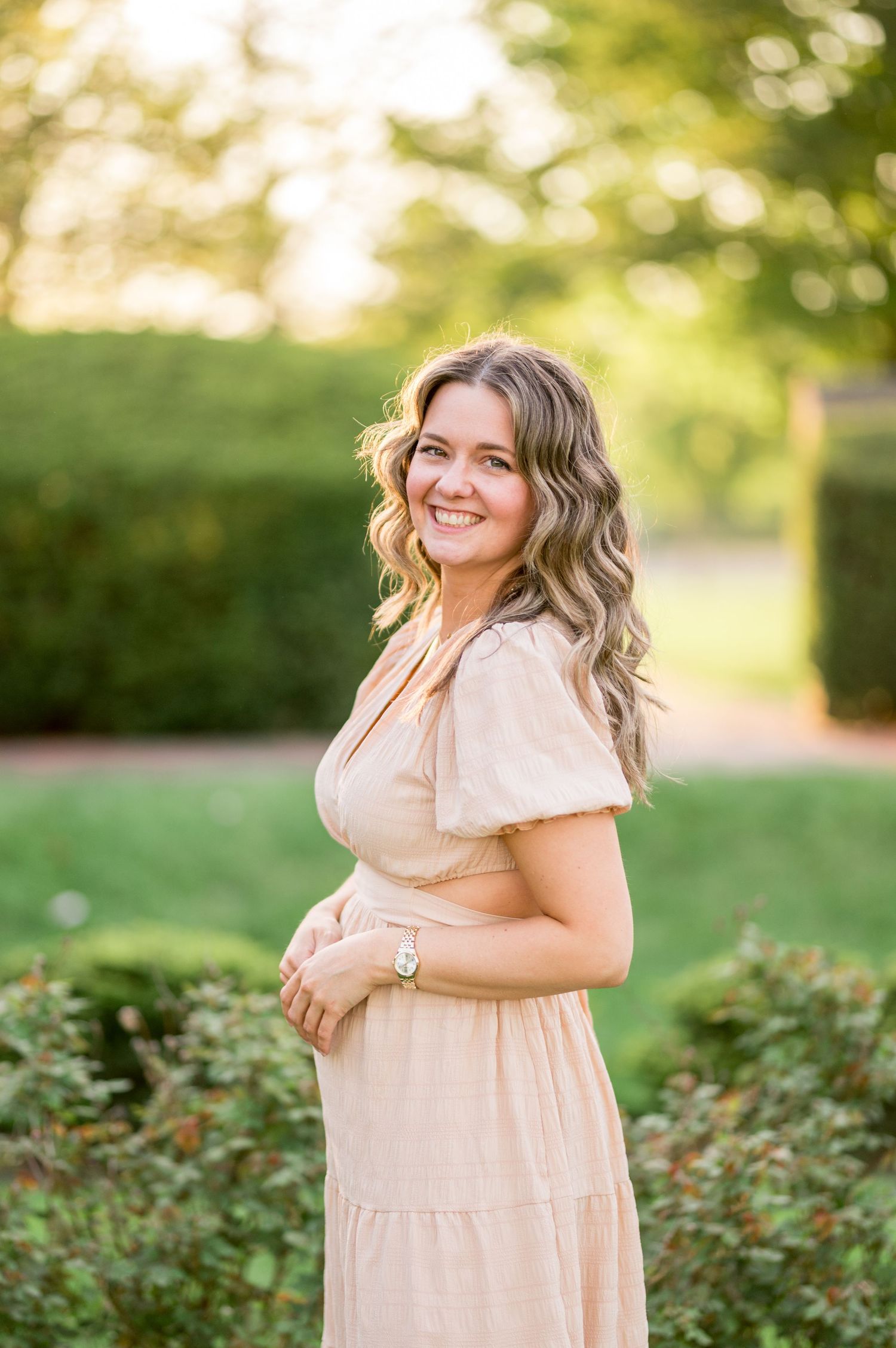 A person in a flowing beige dress poses outdoors in a garden with soft golden sunlight and bokeh in the background.
