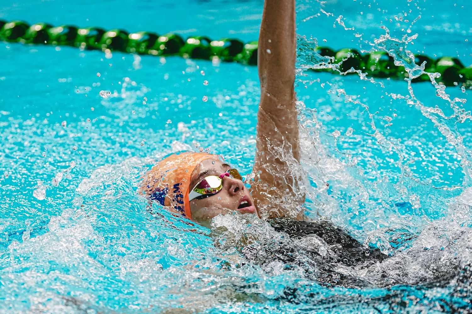 Swimmer performing backstroke in a bright blue pool with splashing water and lane dividers.