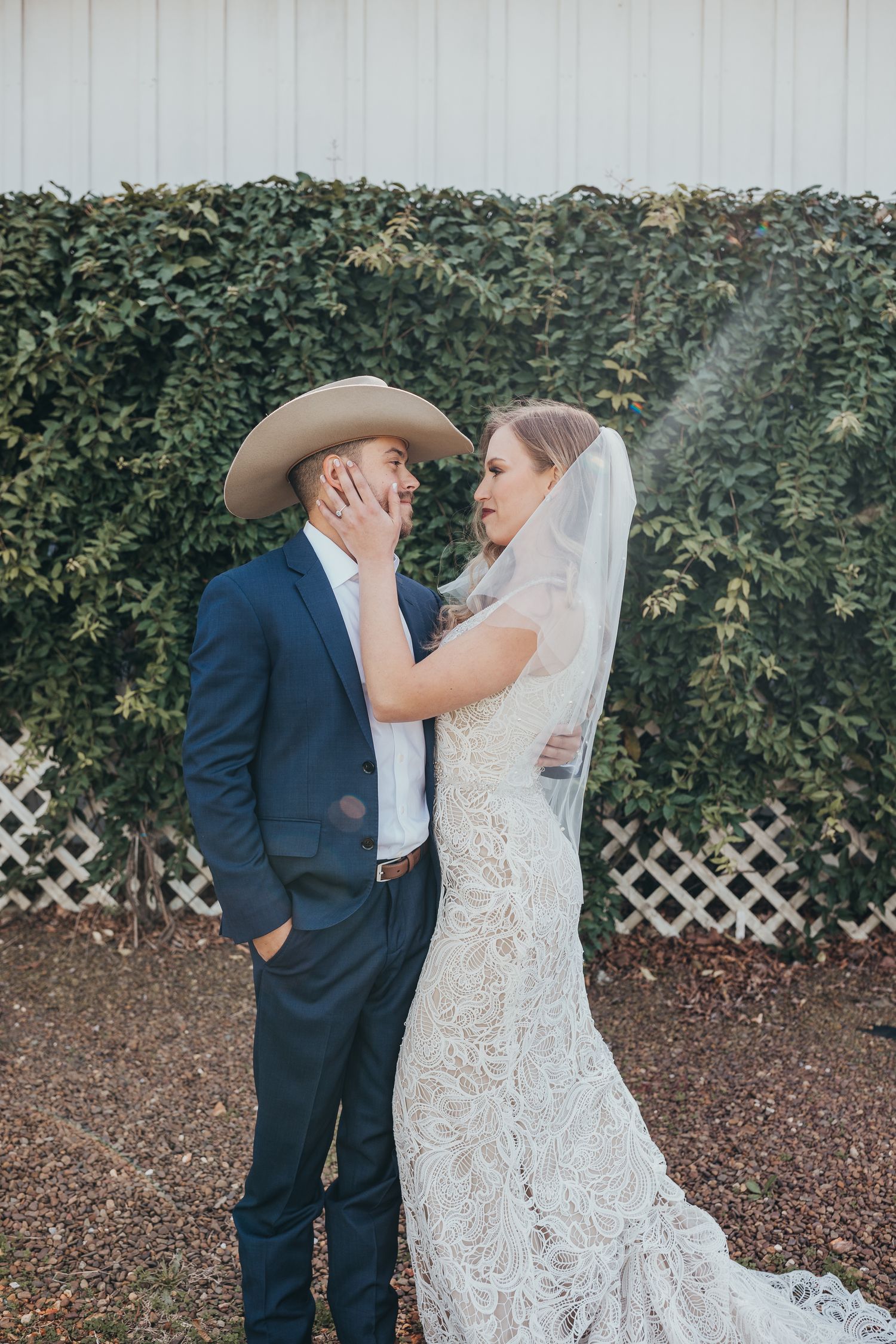 Romantic wedding moment captured in front of a green hedge with cowboy hat and lace gown.