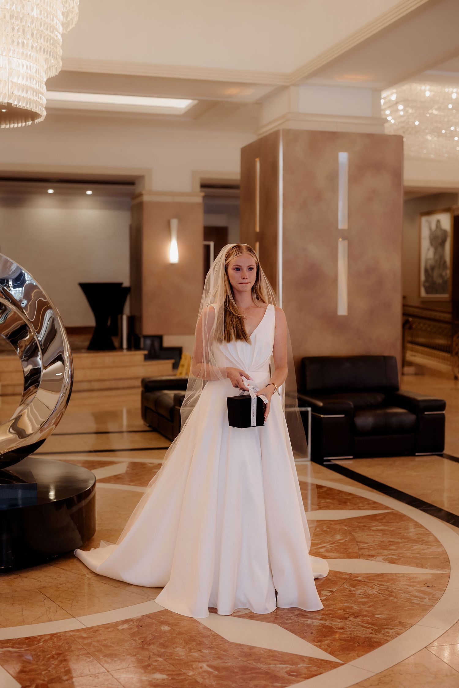 A bride stands in a luxurious hotel lobby wearing a flowing white wedding gown and veil under elegant crystal chandeliers.