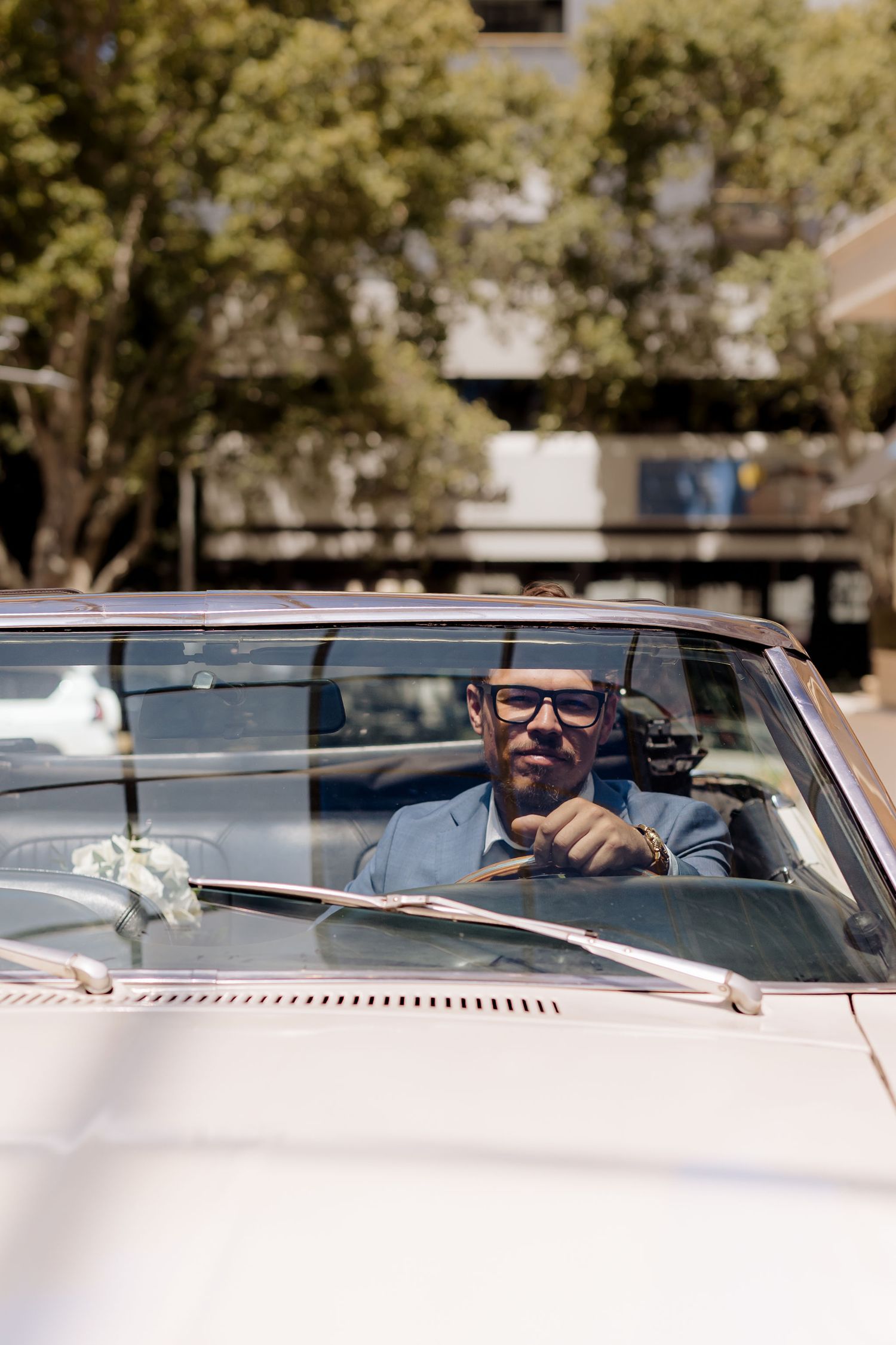 A person wearing sunglasses smiles while sitting in the driver's seat of a classic convertible car on a sunny day.