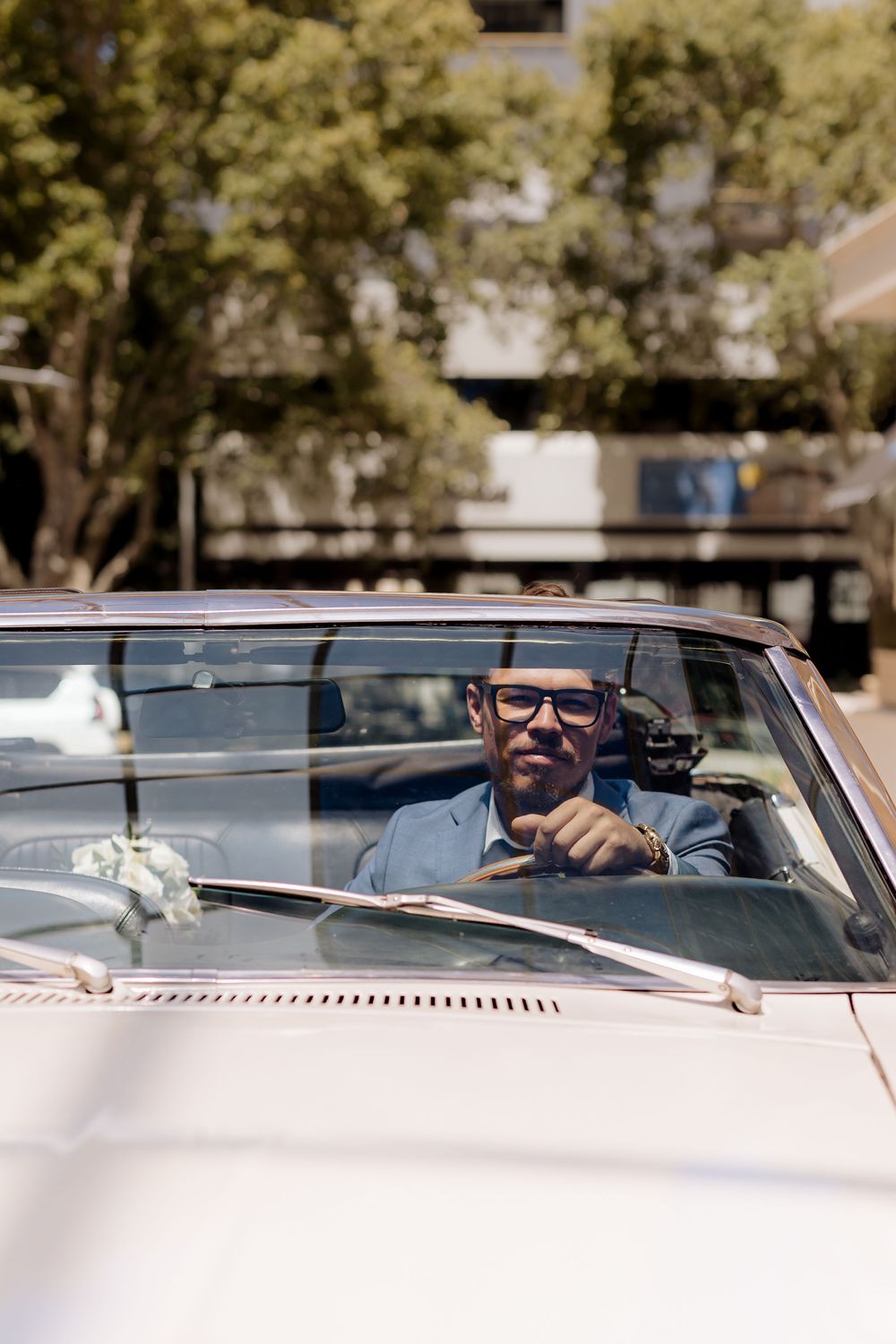 A person wearing sunglasses smiles while sitting in the driver's seat of a classic convertible car on a sunny day.