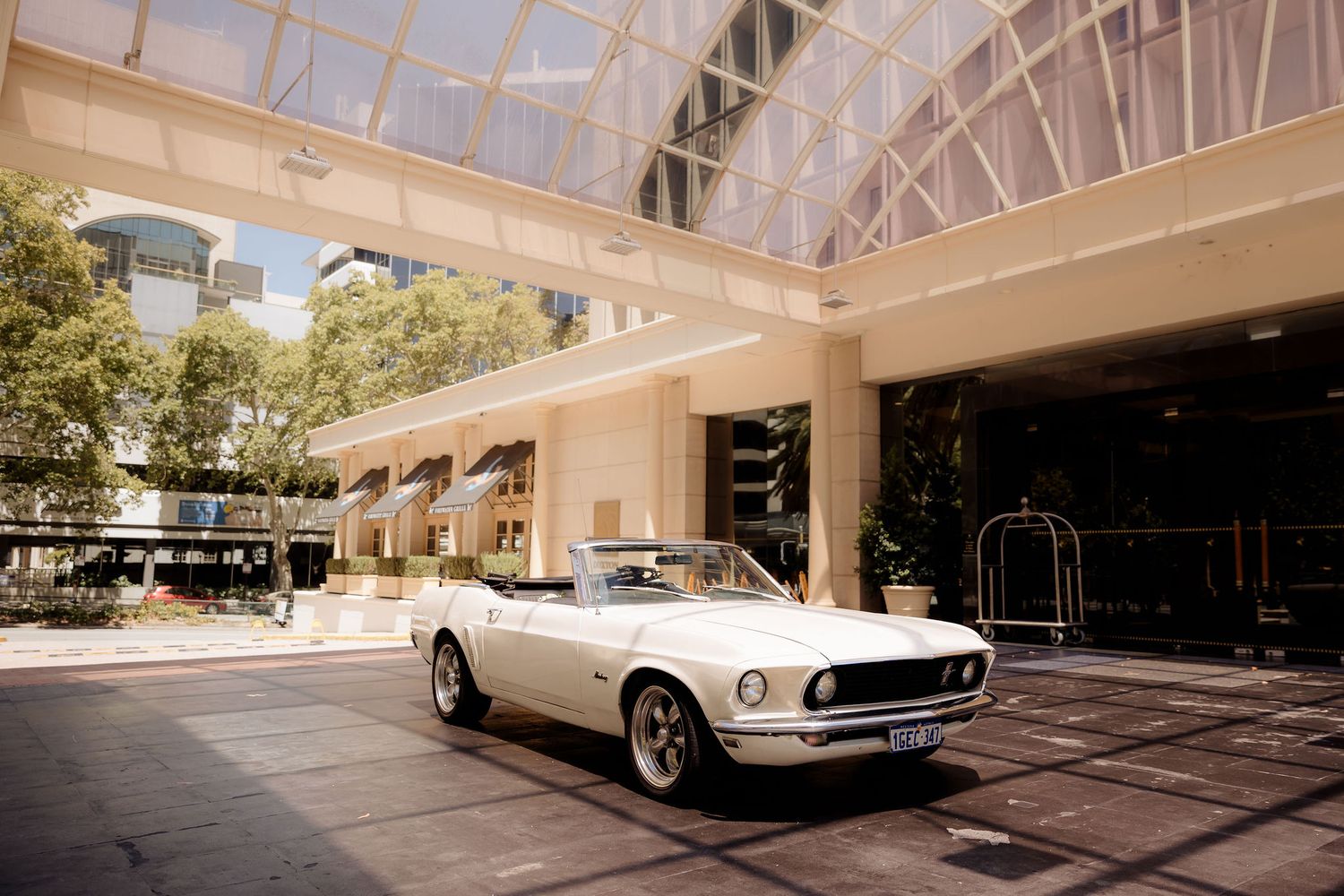 A vintage white Ford Mustang convertible parked under a glass-roofed entrance of an upscale building.