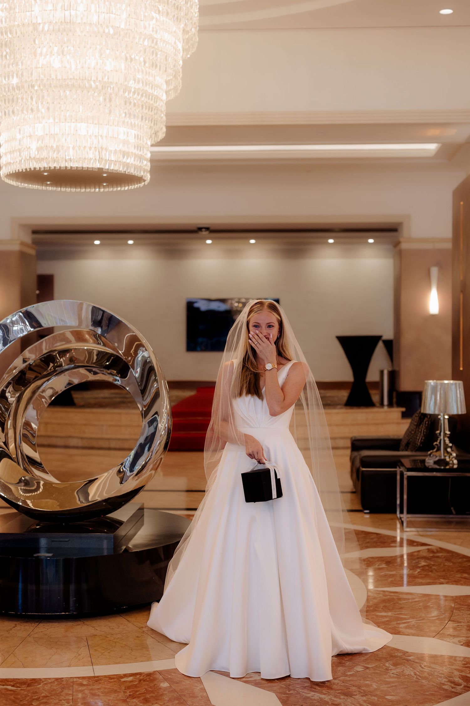 A bride in an elegant white ball gown and veil poses in a luxurious hotel lobby with crystal chandelier and modern decor.