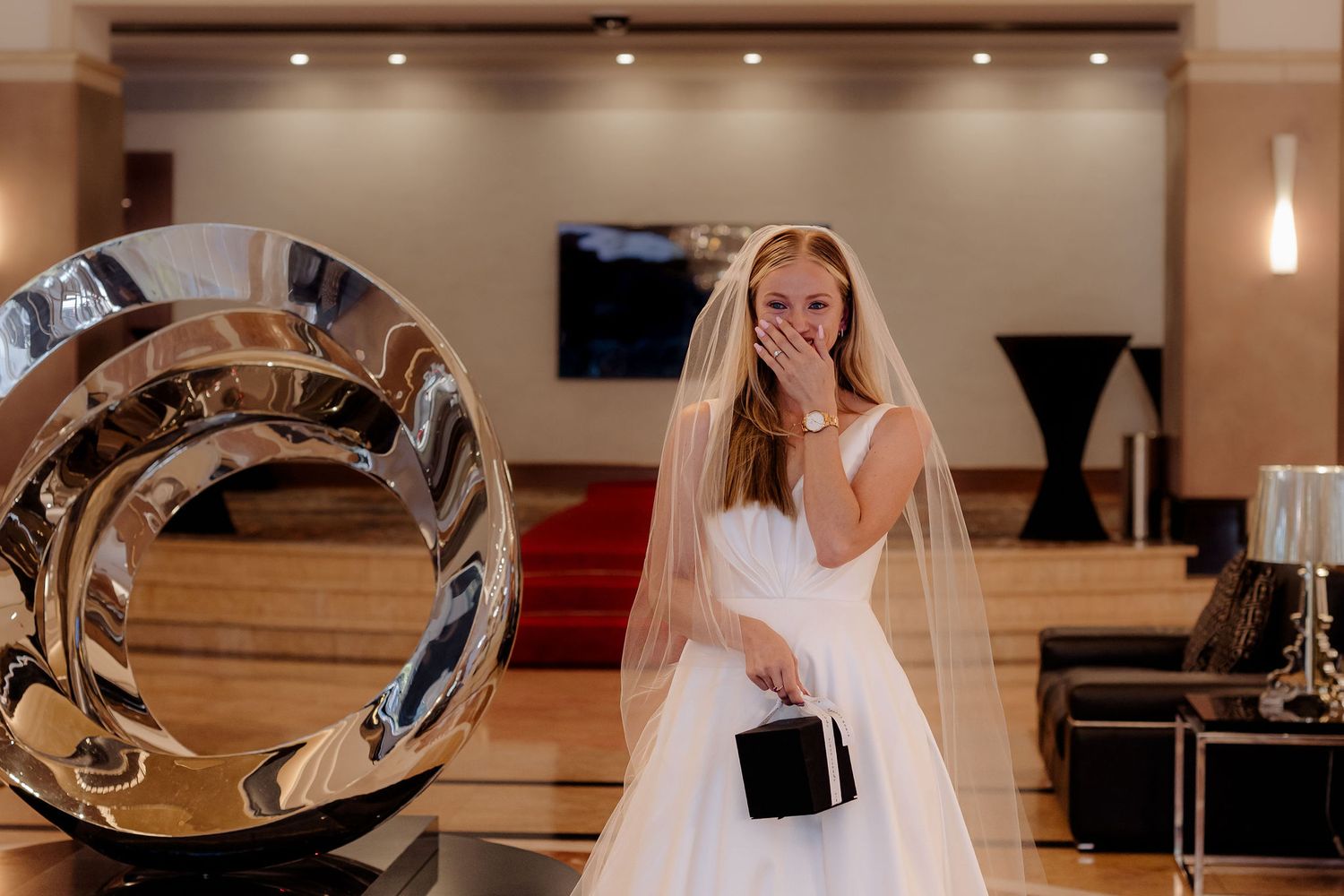 A bride in a white wedding gown stands smiling in an elegant room with modern decor and wood flooring.