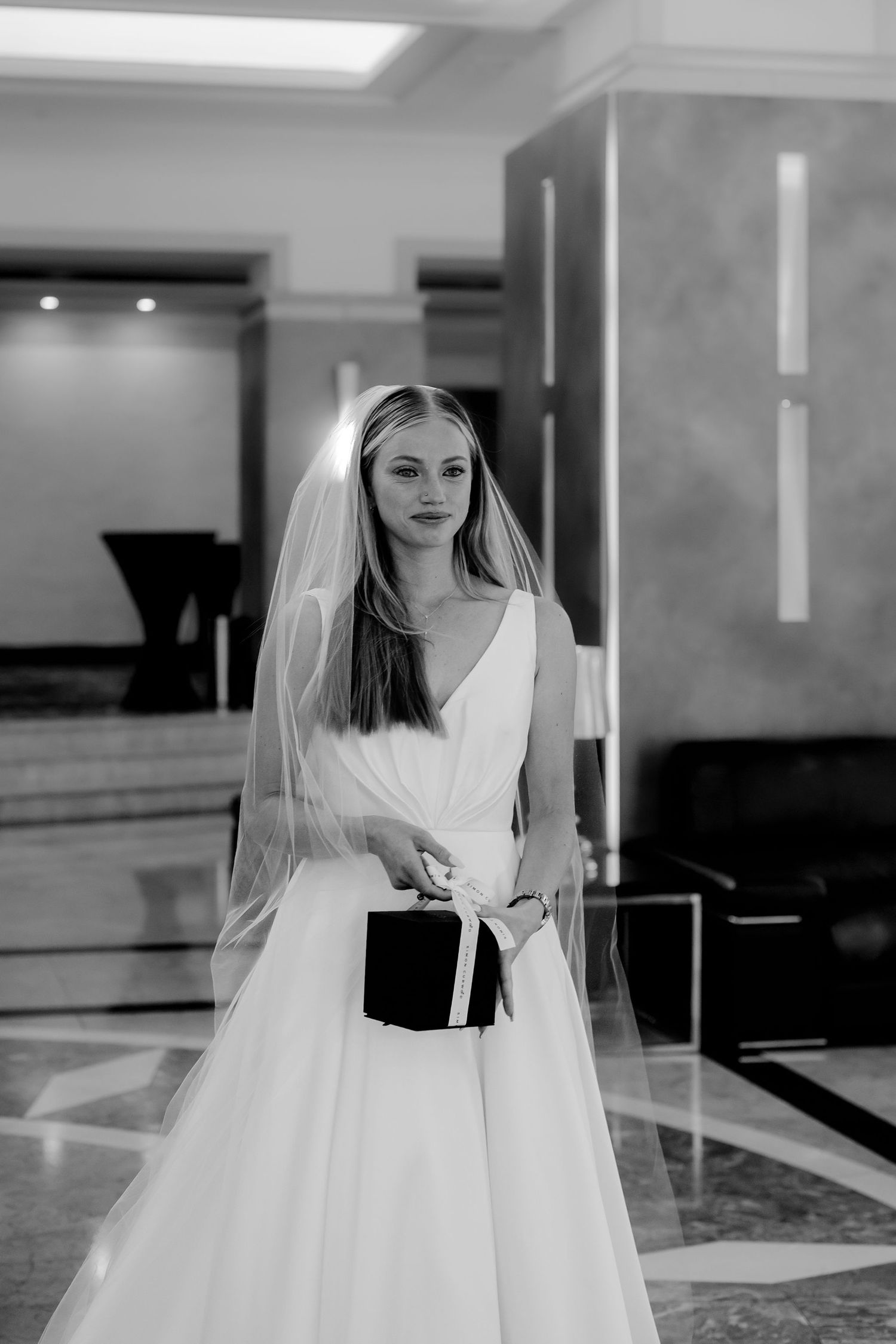 Black and white photograph of a bride in an elegant white gown holding a small black box in a modern interior space.
