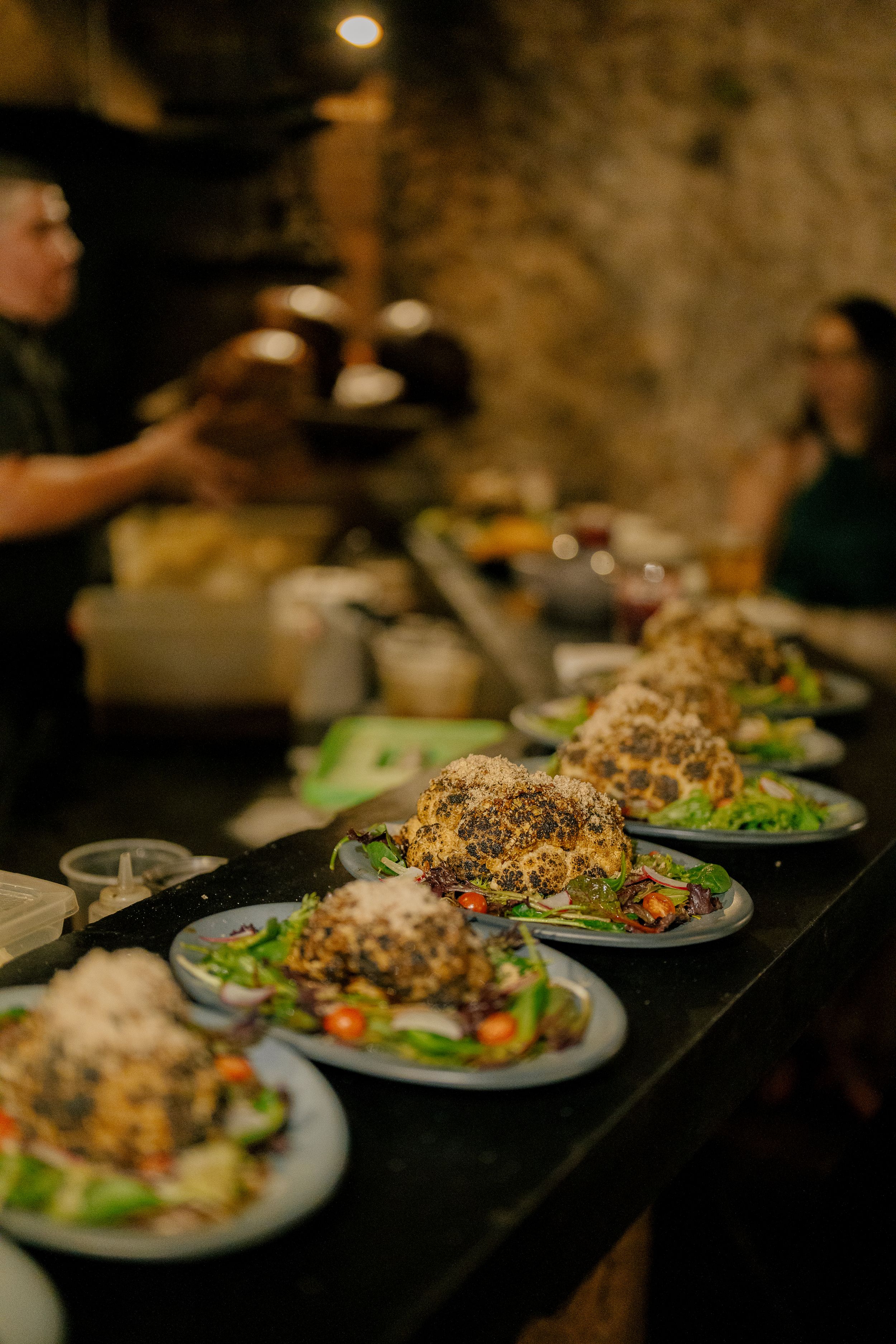 Several plated salads topped with crispy garnish line up on a dark counter in a dimly lit restaurant setting.