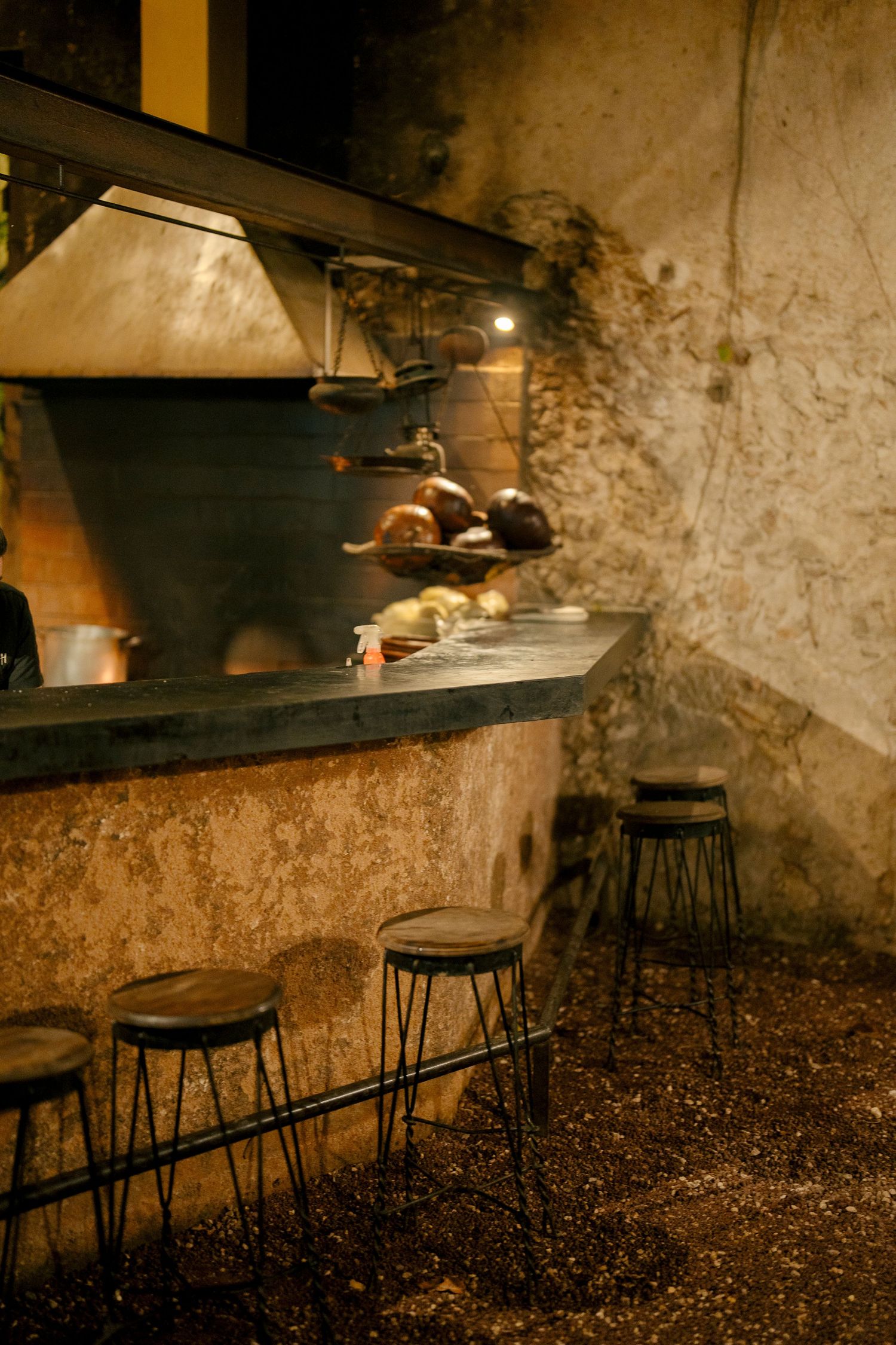 Rustic stone kitchen interior with wood-fired oven and metal bar stools along a dark countertop in moody lighting.