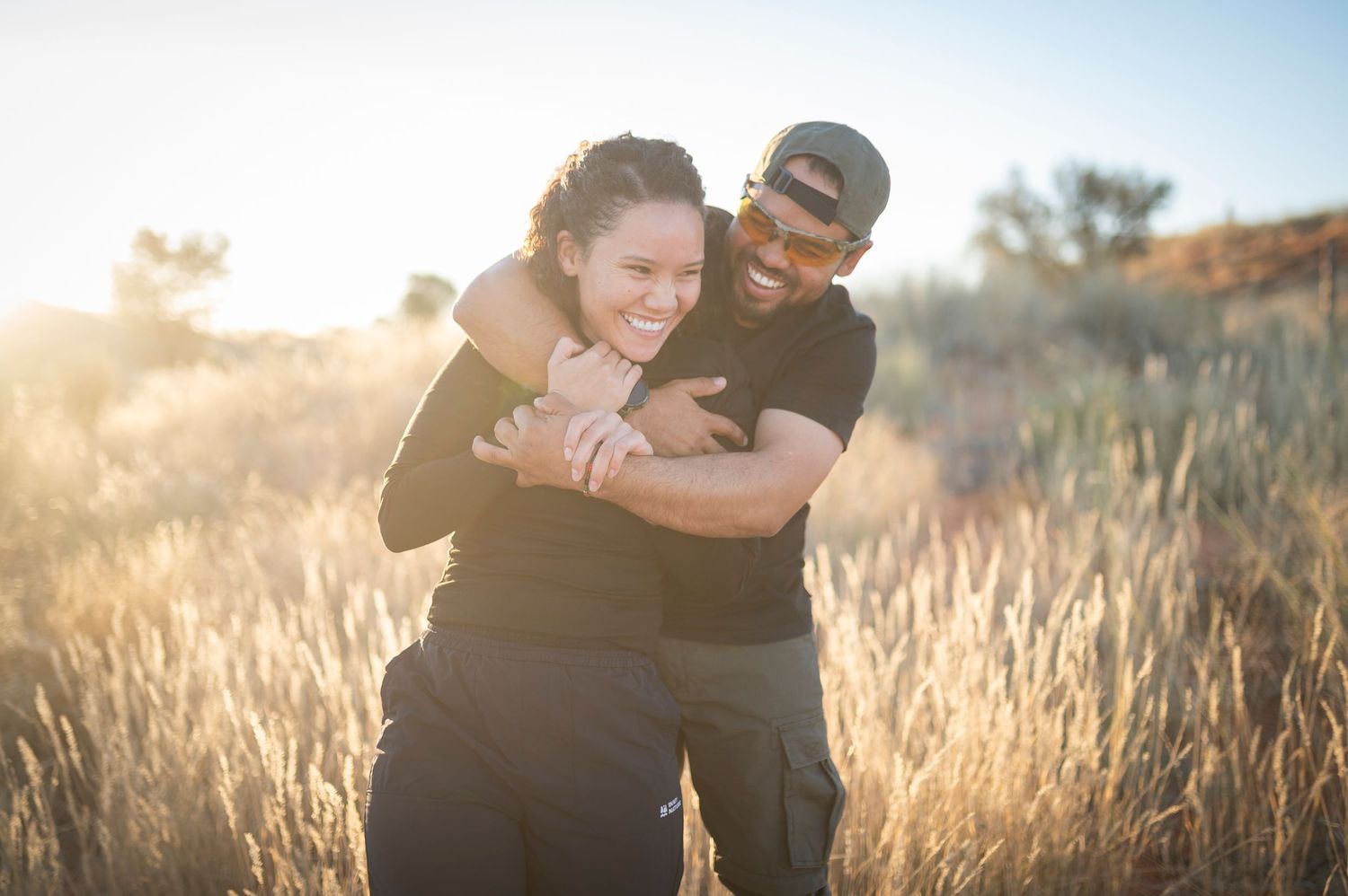 A couple embraces and laughs together in a sunlit field of tall grass during golden hour.