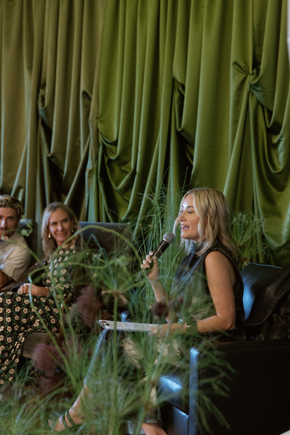 People seated in a stylish venue with lush green curtains and plants creating an elegant ambiance in warm lighting.