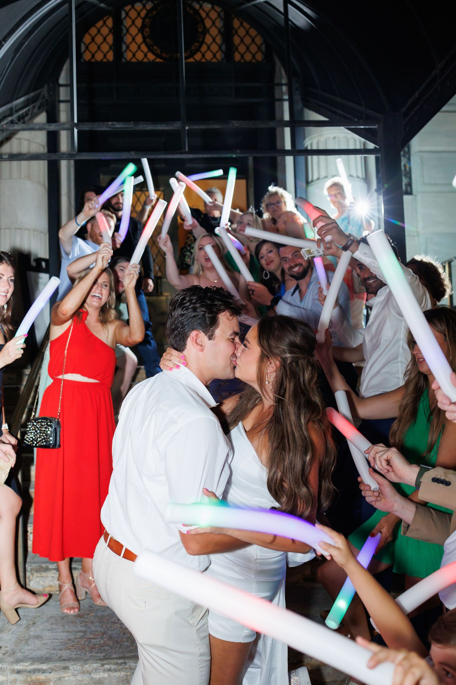 Wedding guests celebrate on the venue entrance holding colorful glow sticks while the couple kisses during their wedding exit