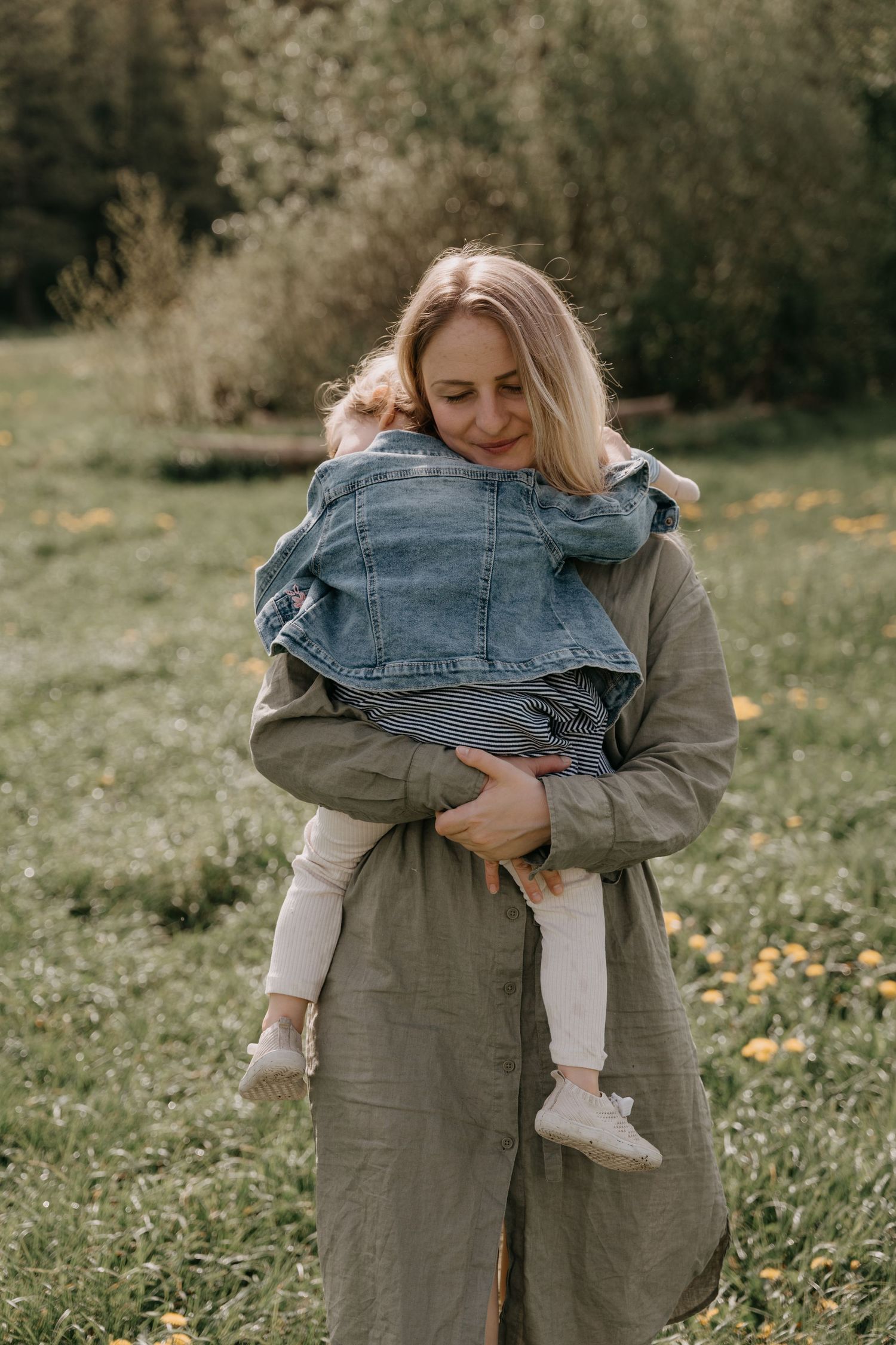 A parent wearing an olive coat holds a small child in denim on a grassy field with blurred trees in the background.