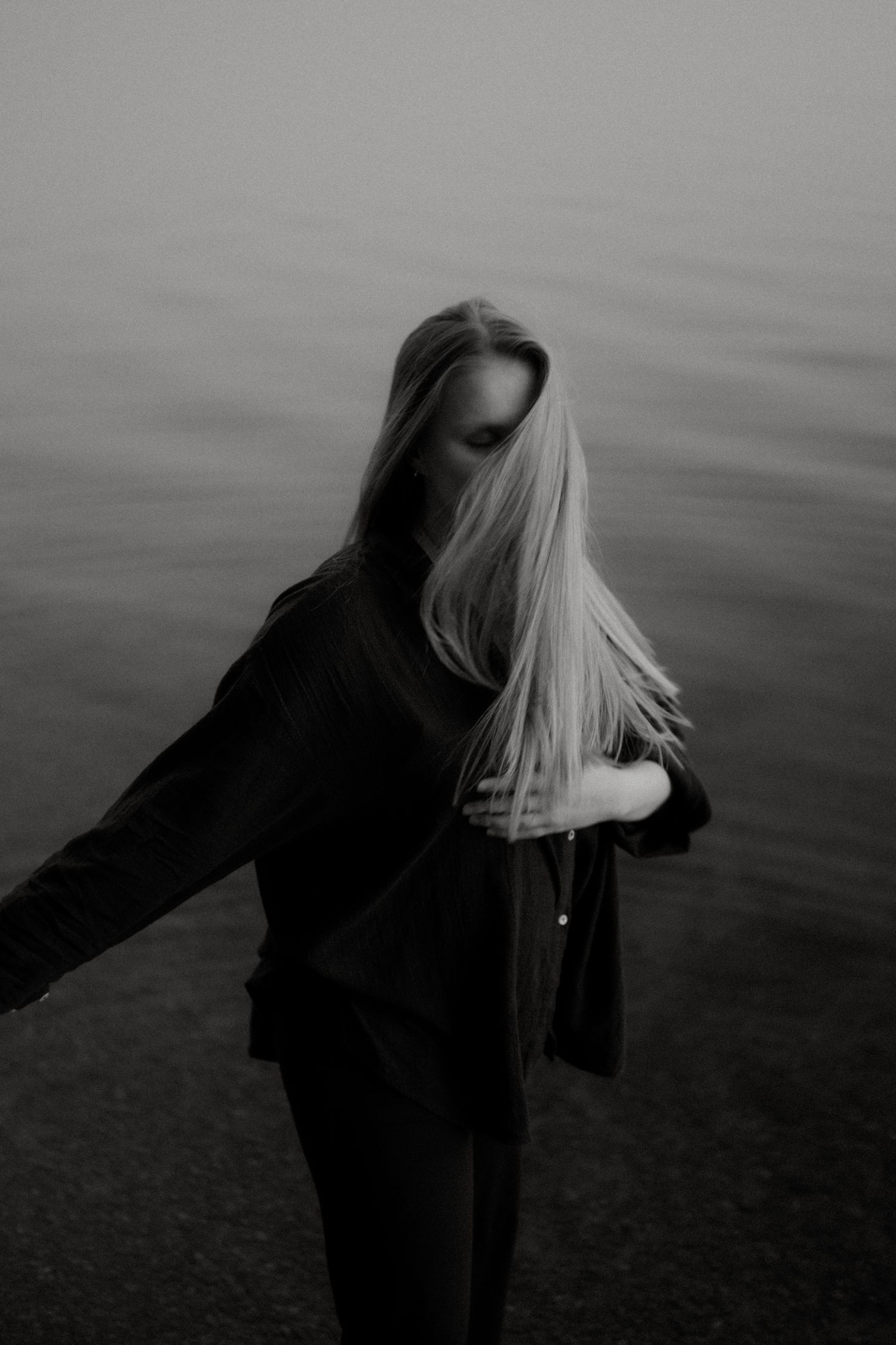 A moody black and white portrait of a figure in dark clothing with flowing blonde hair against a shadowy background.