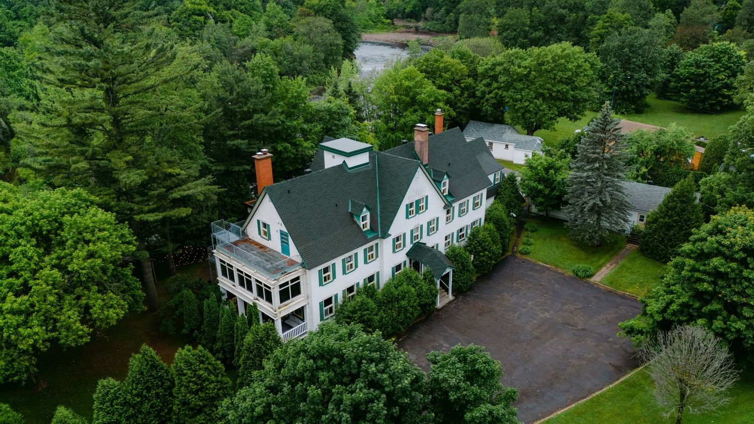 Aerial view of a white colonial mansion with dark gray roof surrounded by lush green trees and circular driveway.