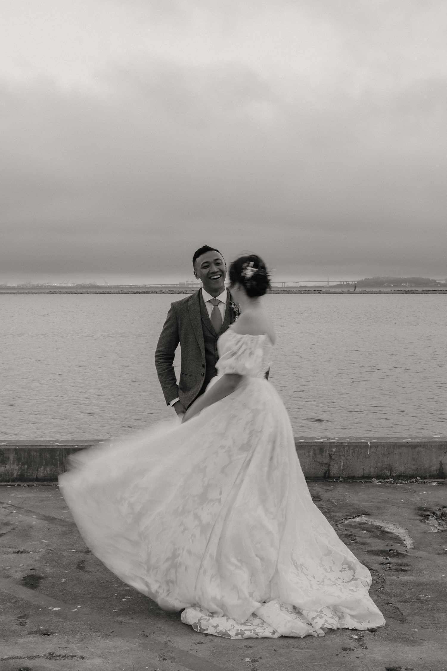 A romantic wedding couple dances and twirls on a beach with water and cloudy skies in the background.