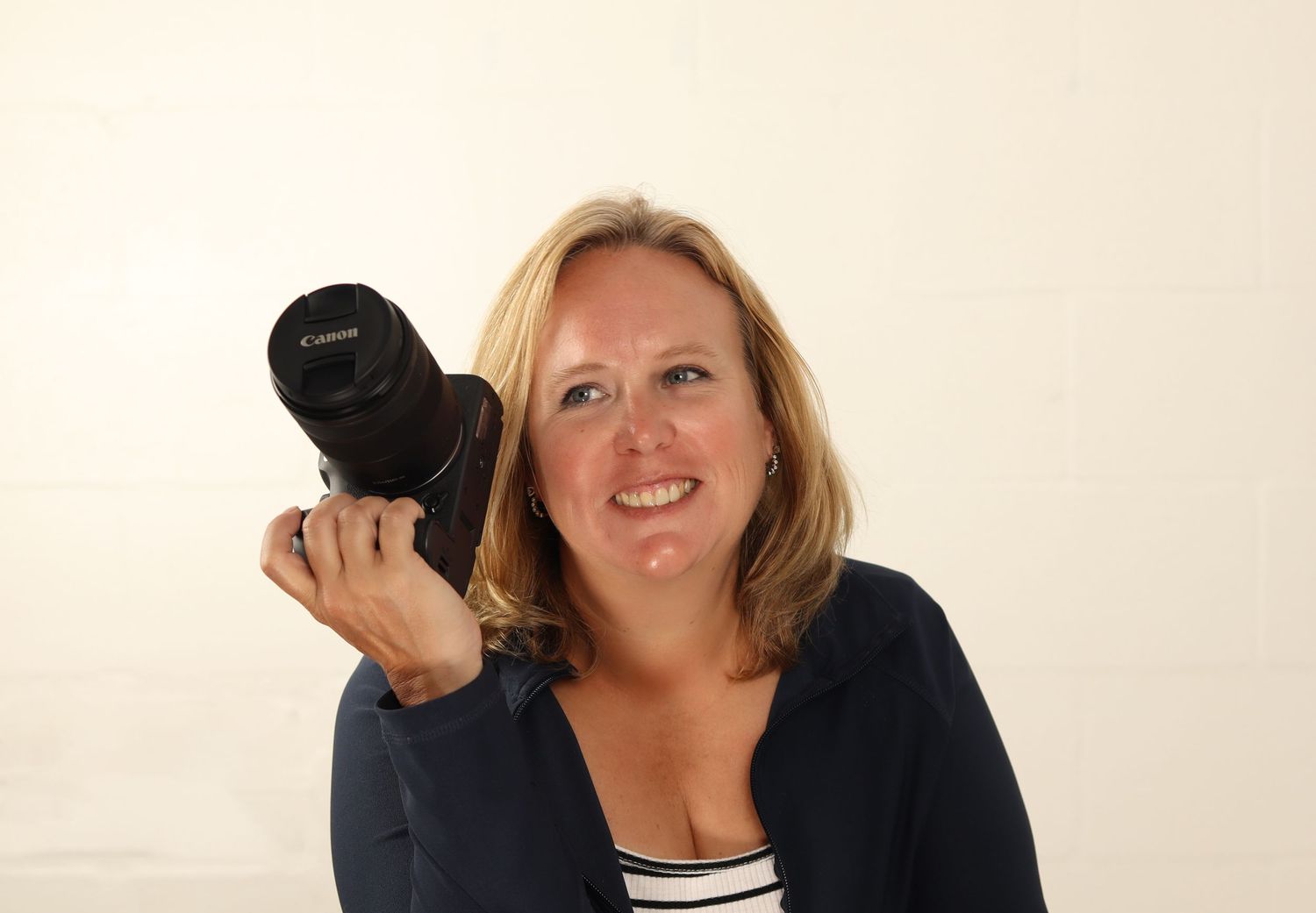 Professional photographer holding a DSLR camera with telephoto lens while wearing navy sweater and striped top.
