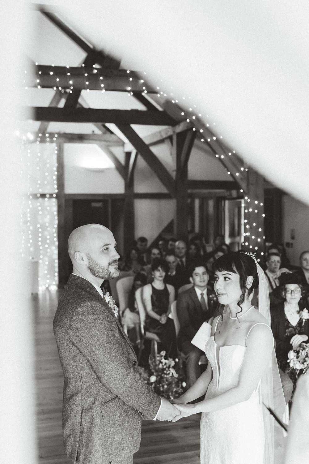 Black and white wedding ceremony photo showing a couple exchanging vows under rustic wooden beams with string lights.