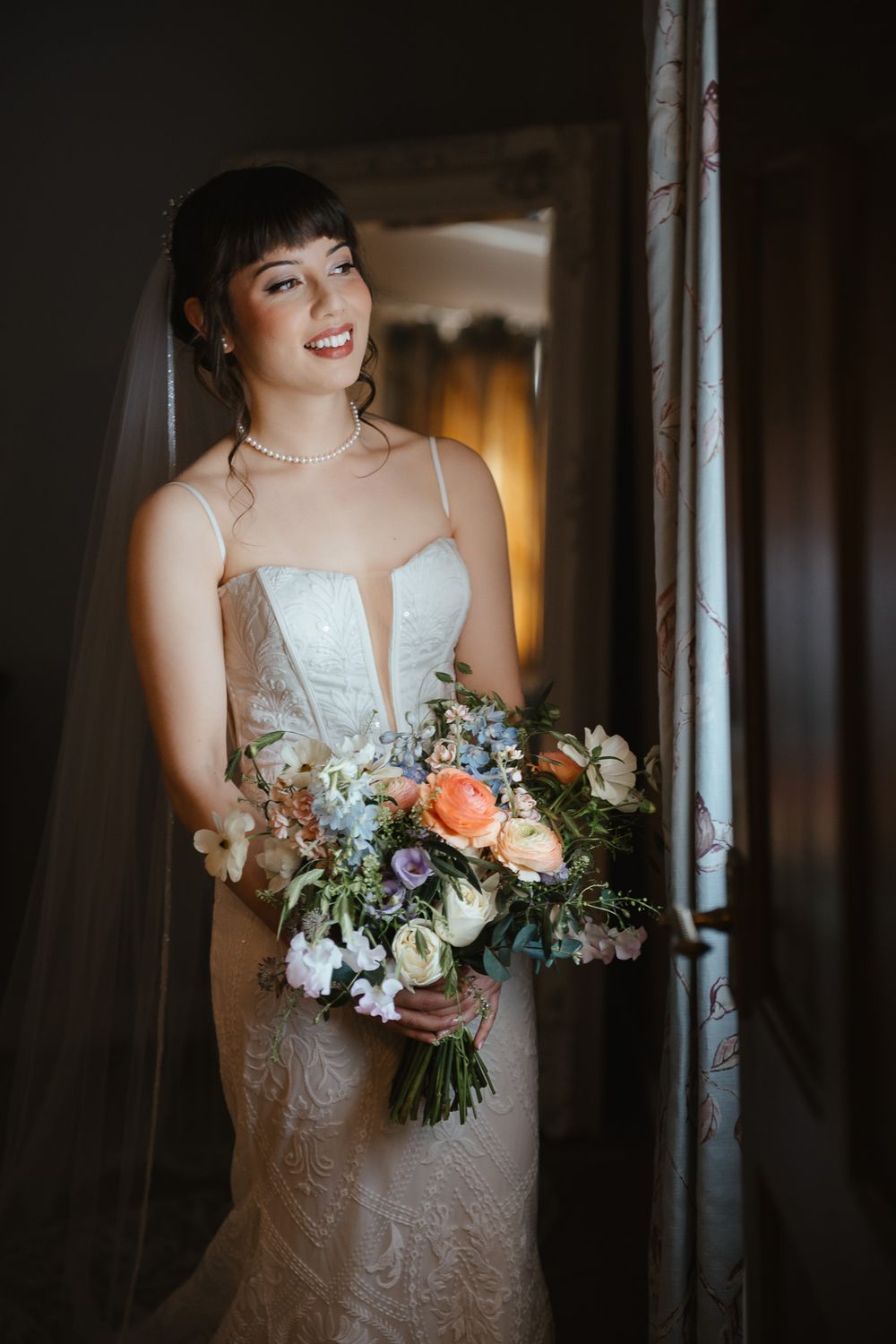 A bride in a sleeveless white gown stands in moody lighting holding a beautiful bouquet with coral and white flowers.
