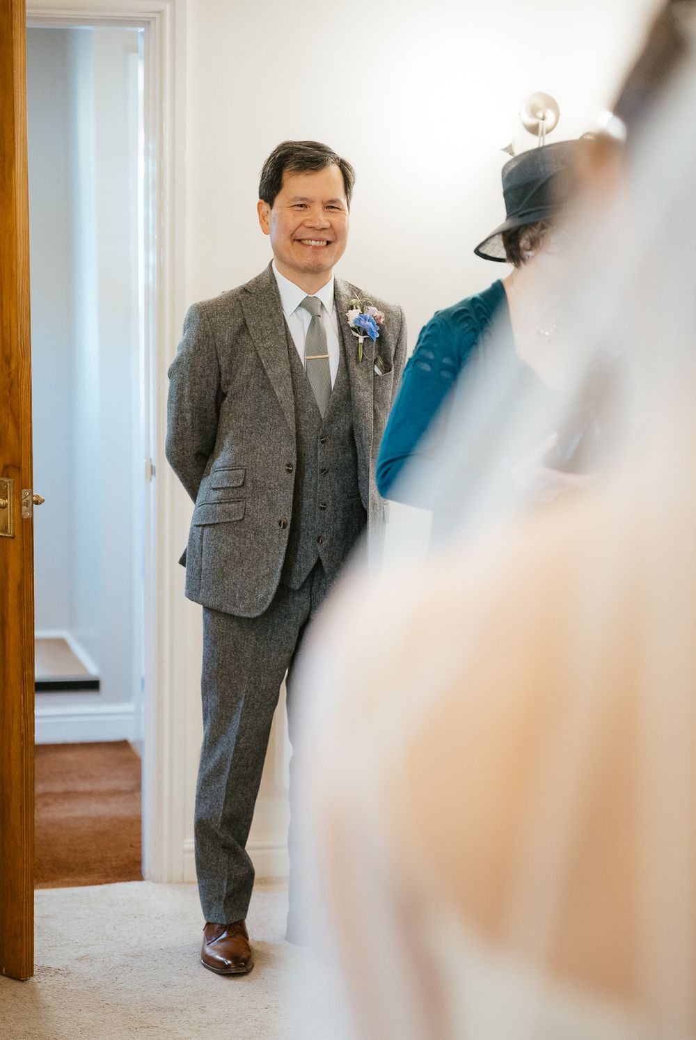 A groom wearing a classic grey three-piece suit smiles while standing in a doorway.