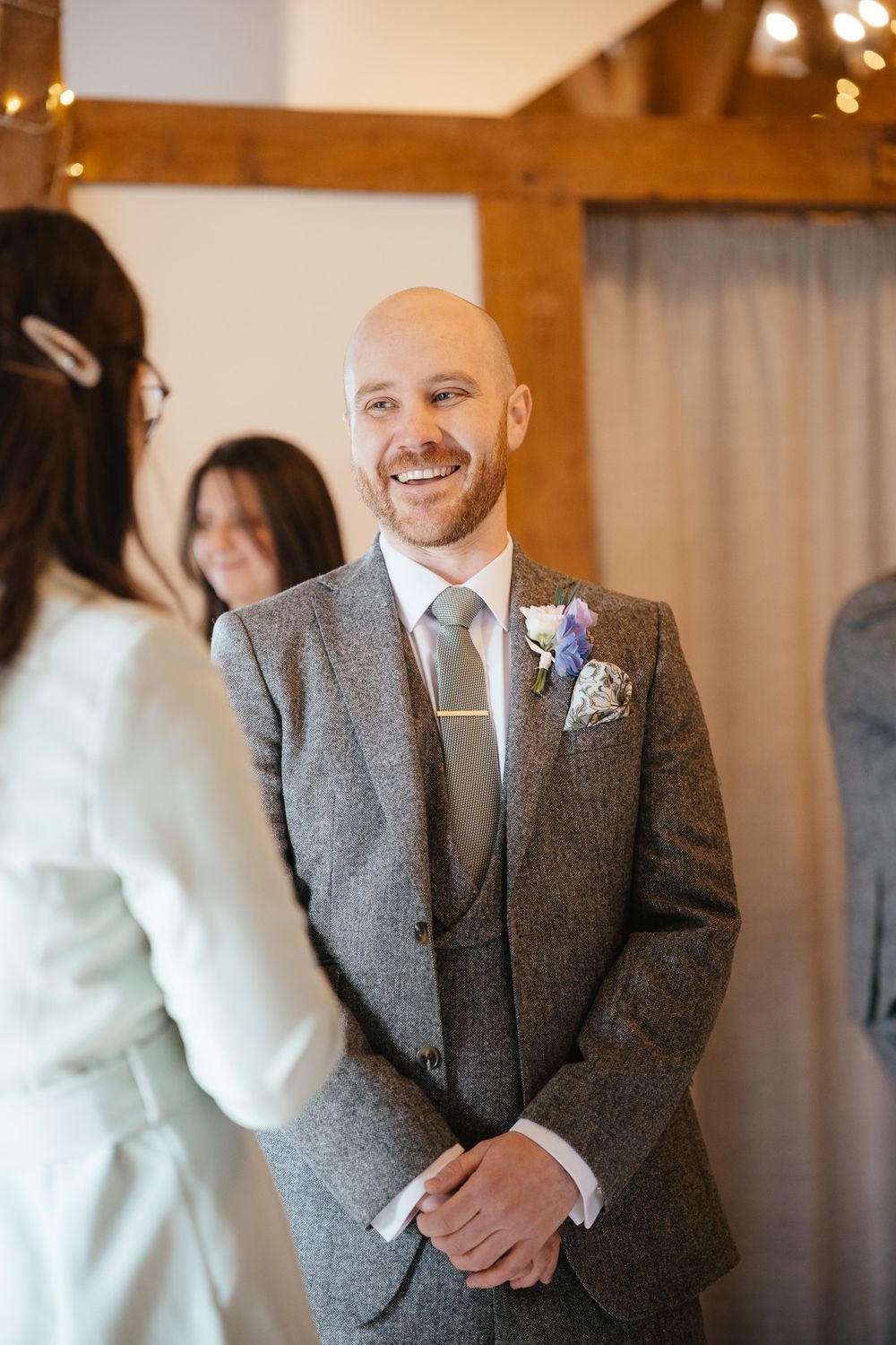 A wedding scene showing someone in a gray tweed suit with a boutonniere smiling warmly during the ceremony.