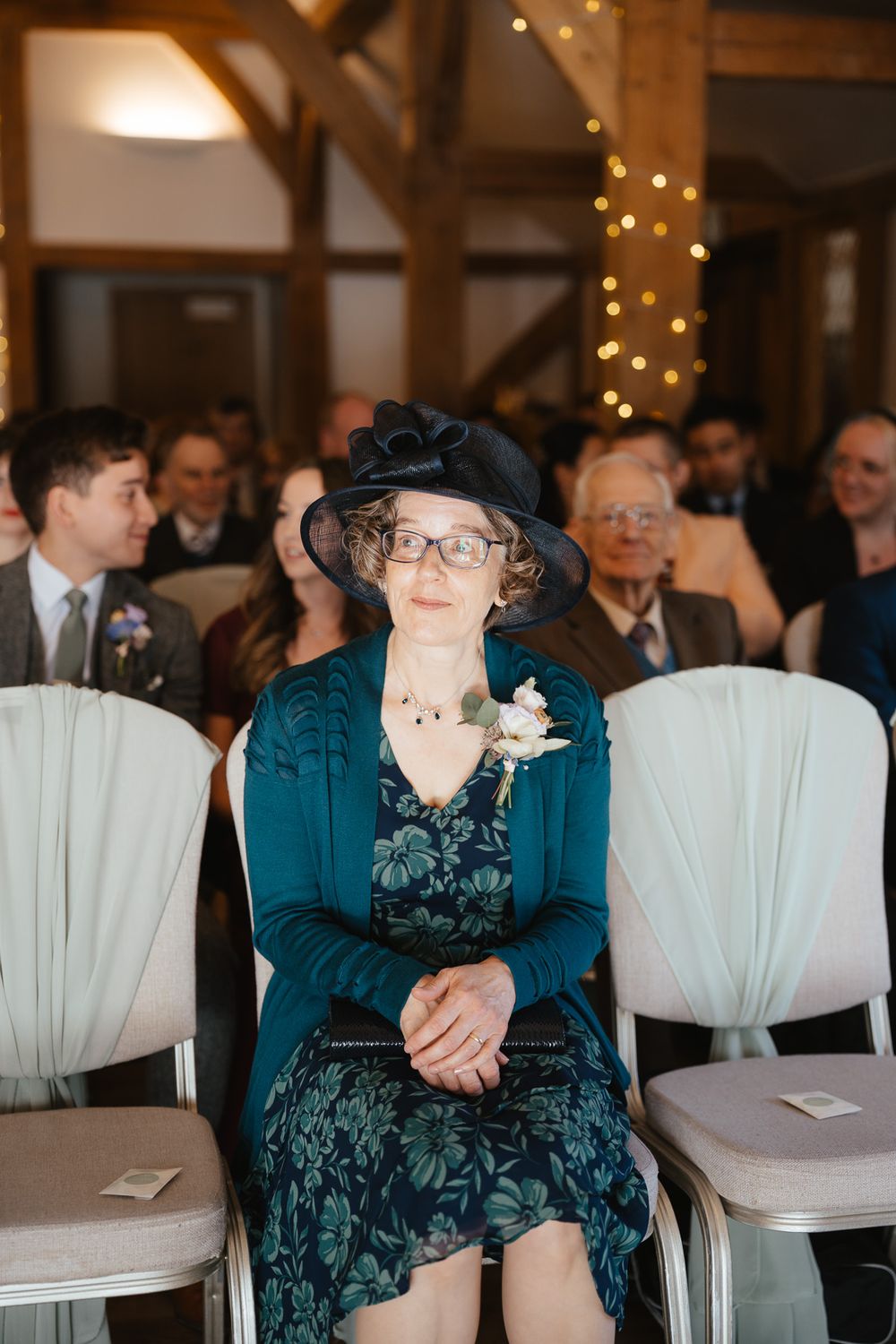 Wedding guests seated in white chairs watch a ceremony in a rustic venue decorated with twinkling lights.