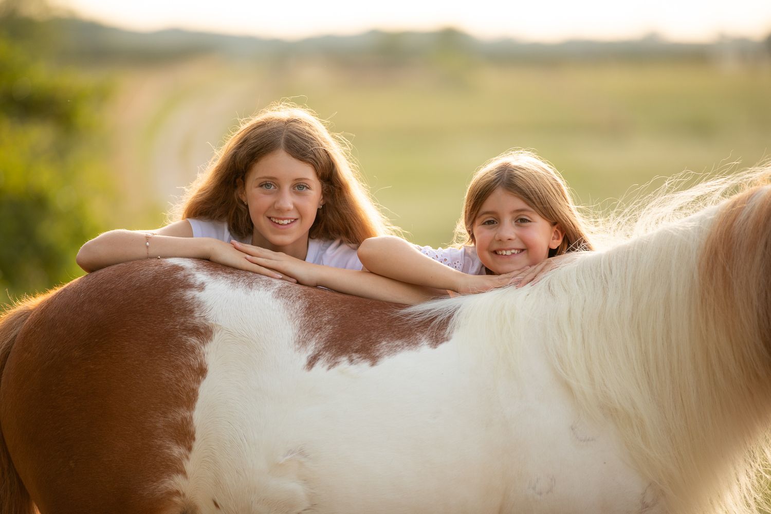 Two joyful riders lean over a white horse's back in a sun-drenched field at golden hour.