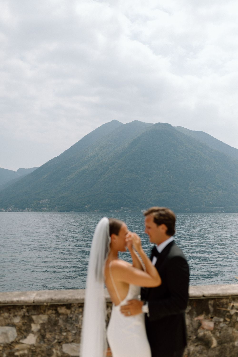 A couple in wedding attire embraces at a lakeside stone wall with mountains rising majestically in the background.