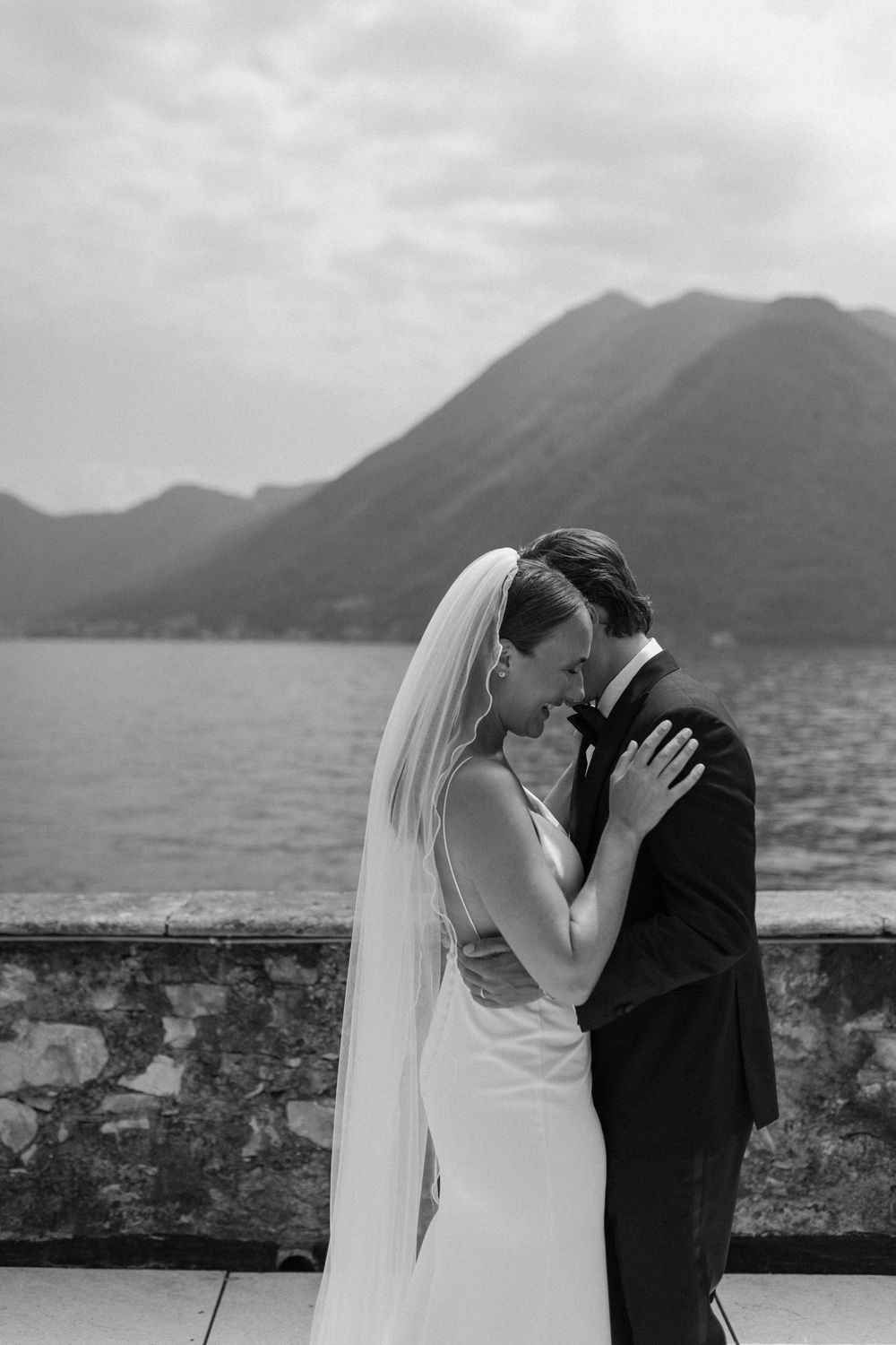 A bride and groom share a romantic kiss along a stone wall overlooking a misty mountain landscape during their wedding.