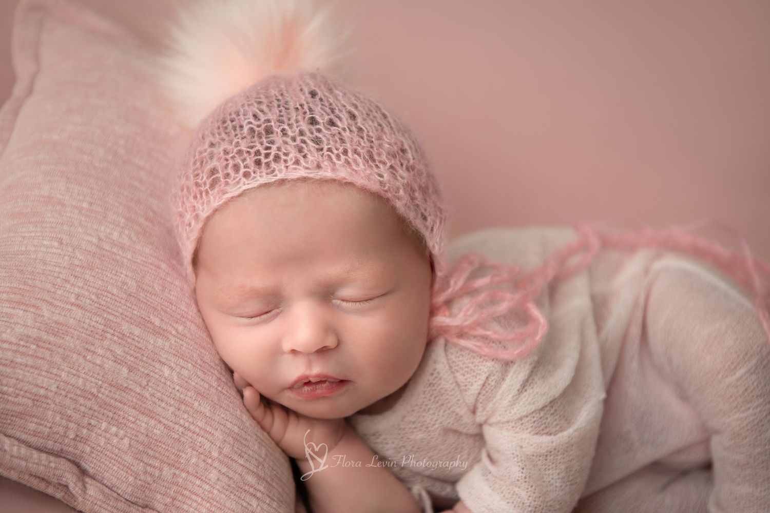 Newborn baby girl sleeping on a pink pillow with hand on her chin in a pink romper and pink heat
