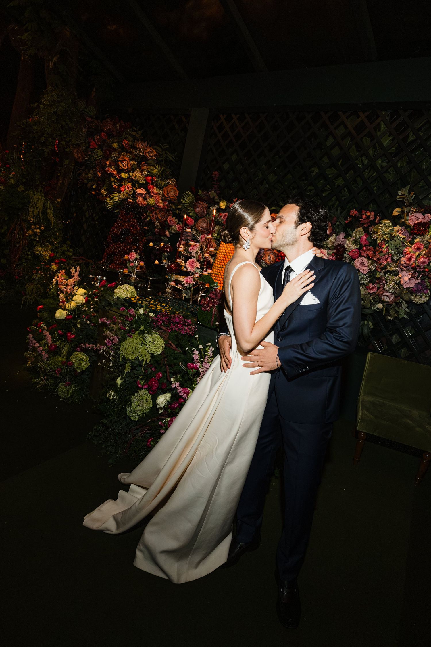 A couple sharing a romantic kiss at night against a backdrop of beautiful pink and red floral arrangements.
