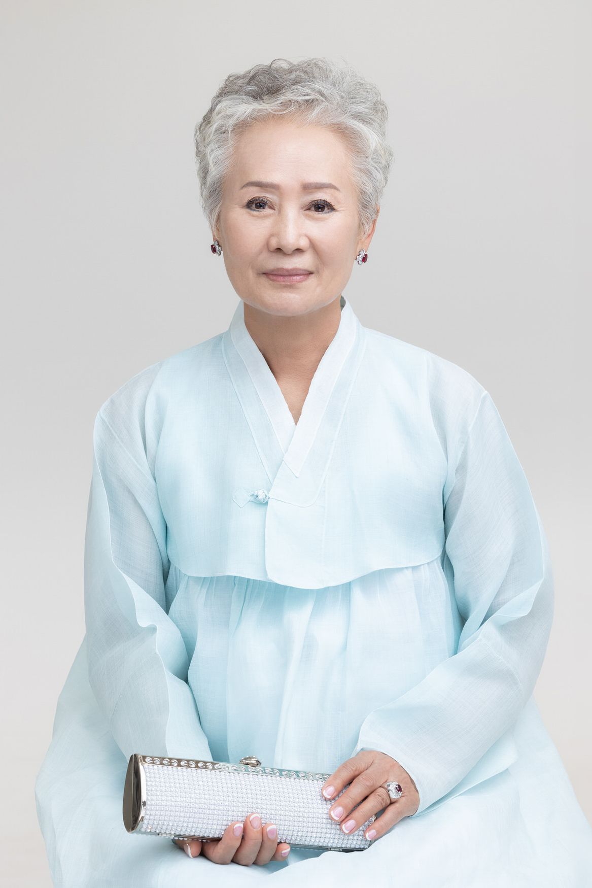 A woman in a light blue hanbok holds a white clutch purse while seated against a gray background.