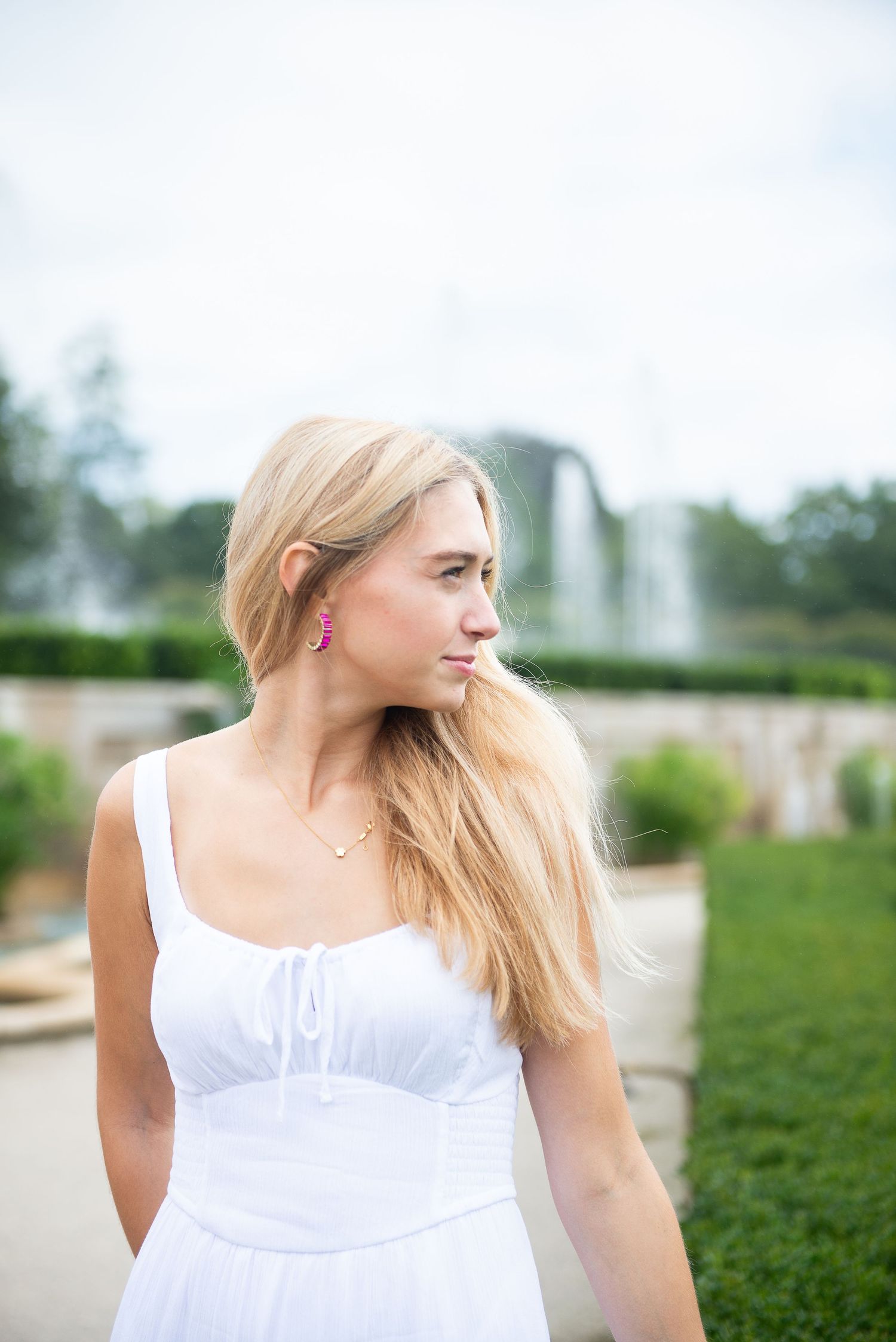A person in a white summer dress stands in a garden with a fountain in the background on a sunny day.