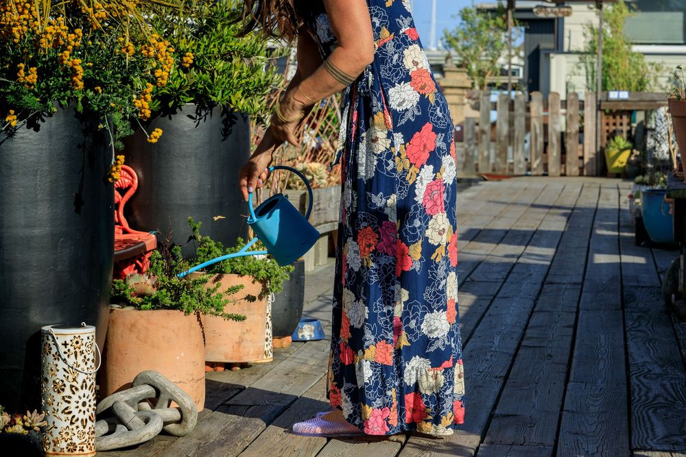 A person in a floral dress walks along a wooden deck pathway lined with potted plants and flowers.
