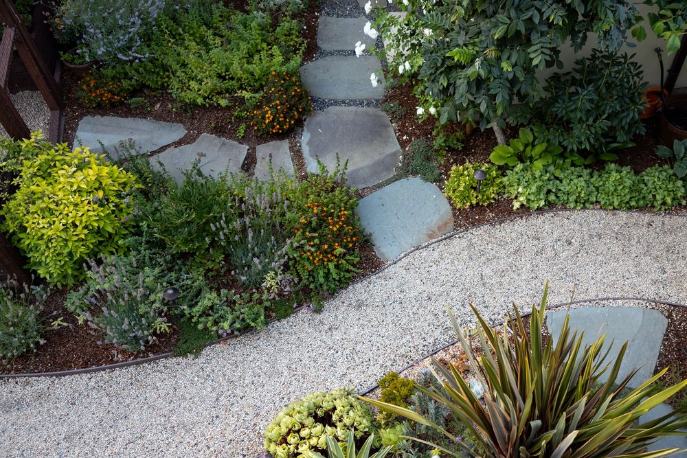 A curved gravel path winds through a modern garden landscape with decorative concrete walls and diverse plantings.