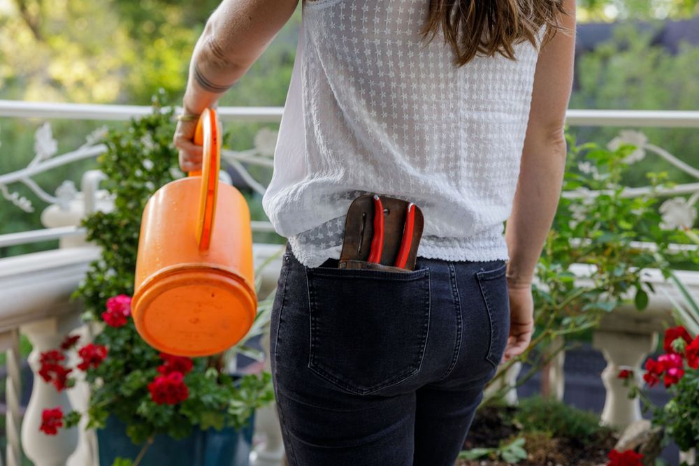 Someone holding a bright orange watering can on a balcony garden with red flowers in the background.