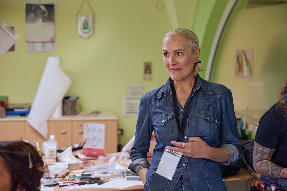 Workshop facilitator wearing denim shirt stands attentively while holding materials in bright green room.