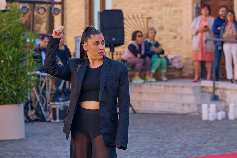 A performer in a black blazer and crop top dances in an outdoor courtyard with spectators in the background.