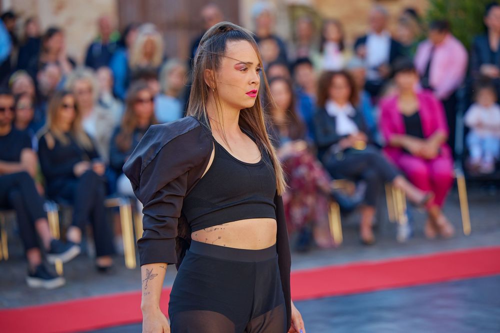 A performer in a black crop top and pants stands confidently on a red carpet with spectators in the background.