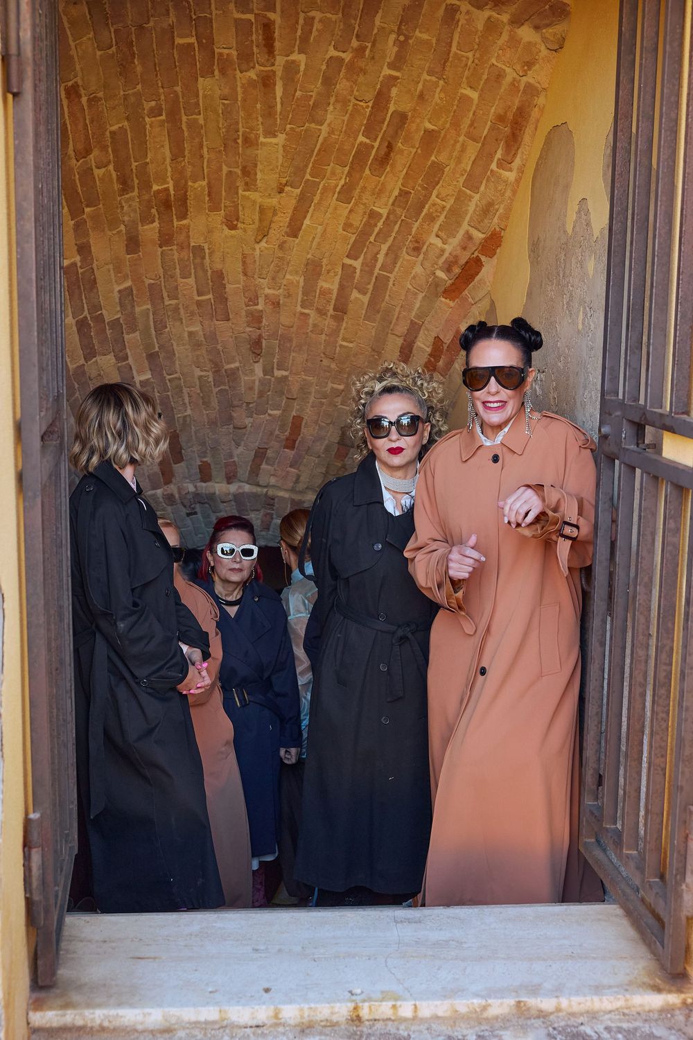 Three figures in long dark coats and one in peach stand in a doorway wearing sunglasses while smiling and laughing.
