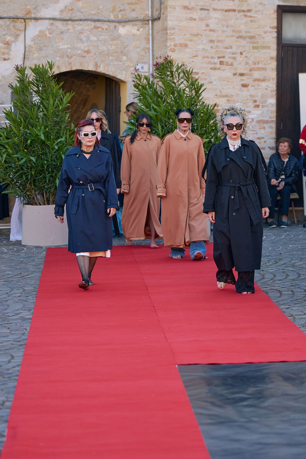 Models walk a red carpet runway wearing long coats and stylish sunglasses during an outdoor fashion event.