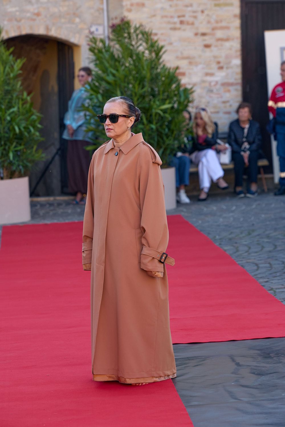 A person wearing sunglasses and a long camel coat poses on a red carpet at an outdoor event.