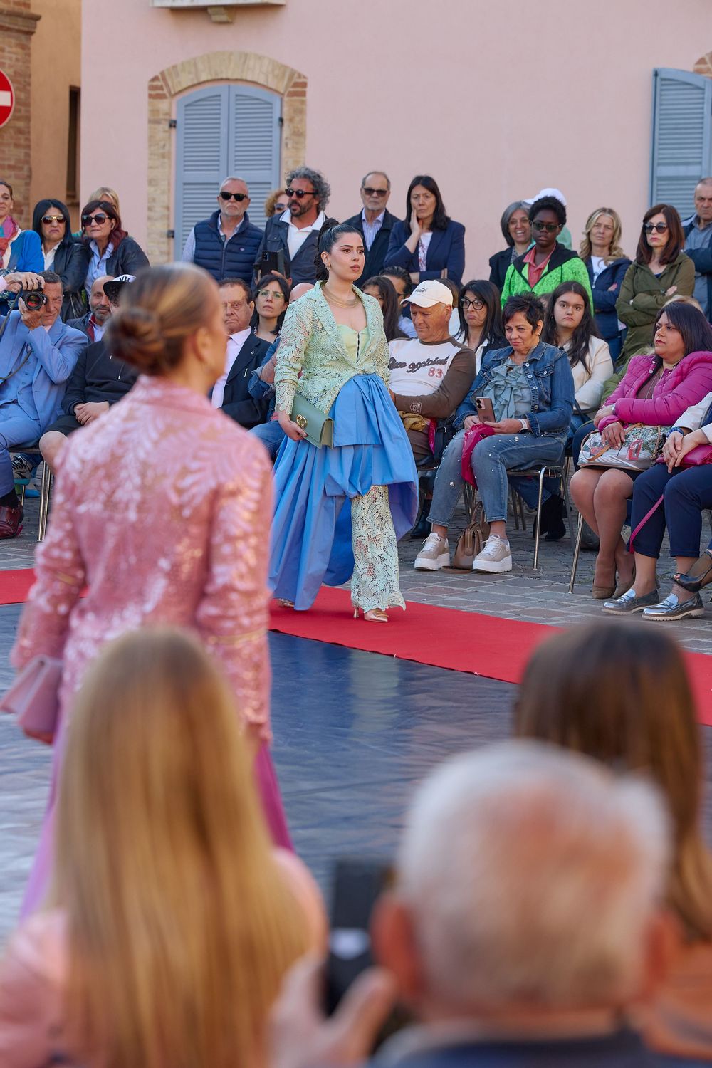 A crowd watches from the sidelines during an outdoor fashion show with models walking down a red carpet runway.