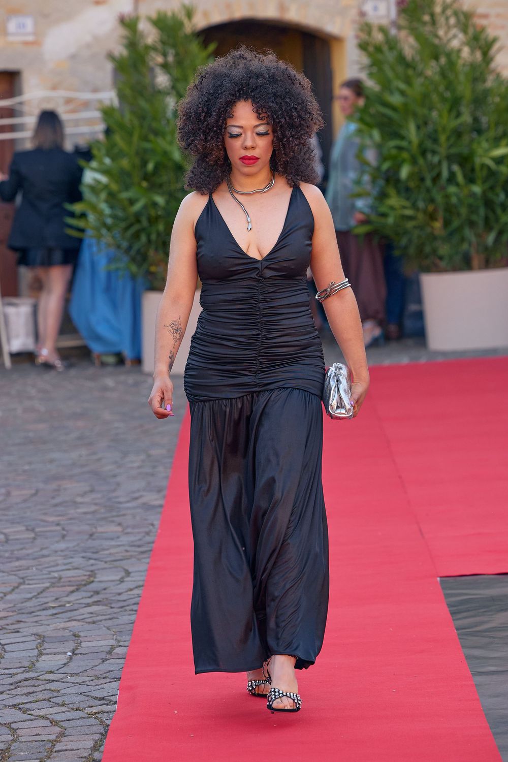 A figure walks down a red carpet in a black formal gown with ruched detailing at a glamorous outdoor event.
