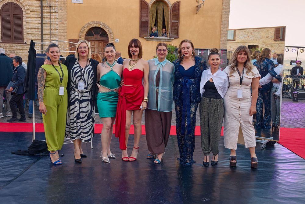 A group of people in colorful outfits stand together on a dark floor mat against a beige wall backdrop.