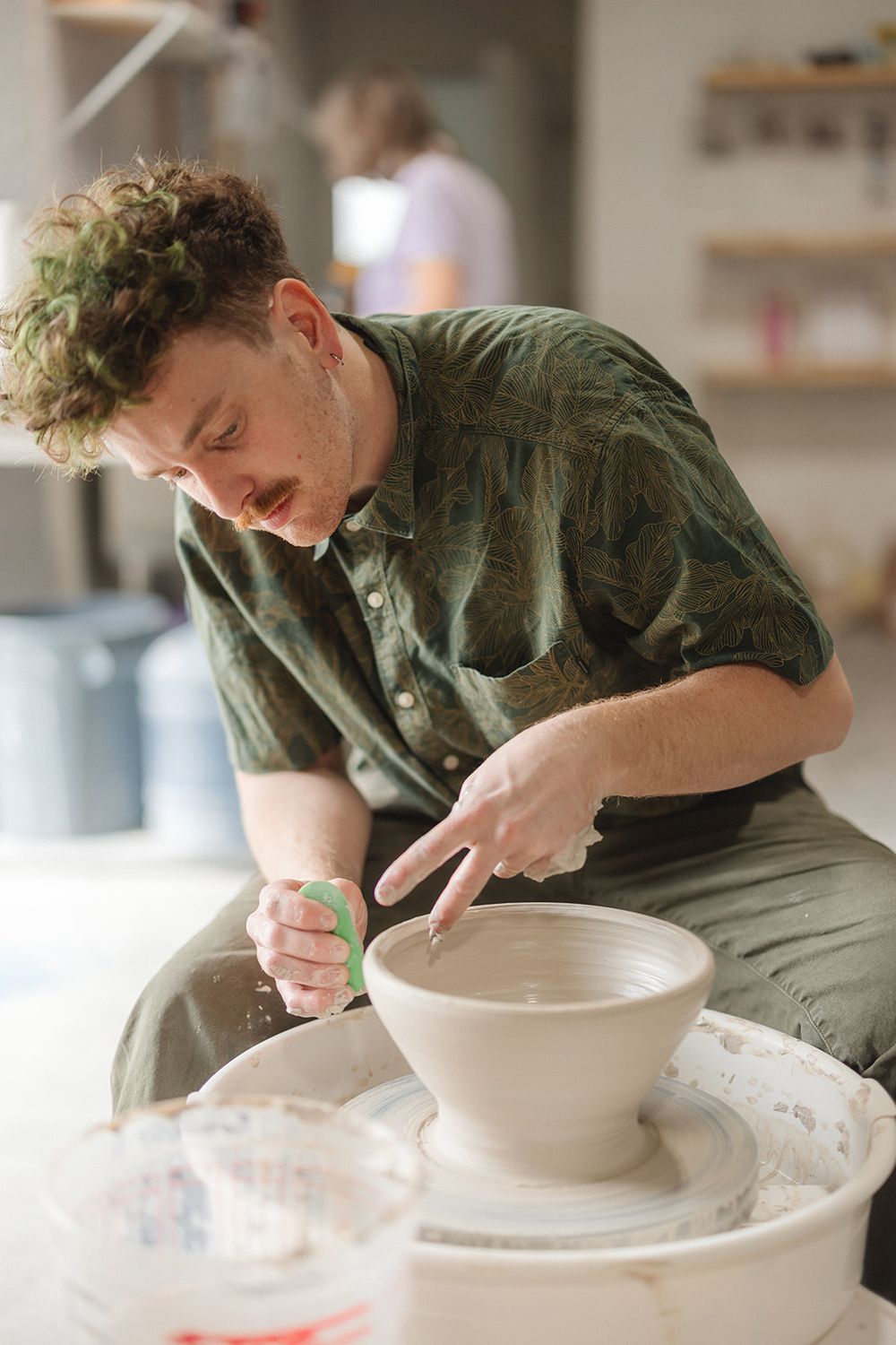 An artist in an olive green shirt shapes clay on a pottery wheel while concentrating intently on their work.