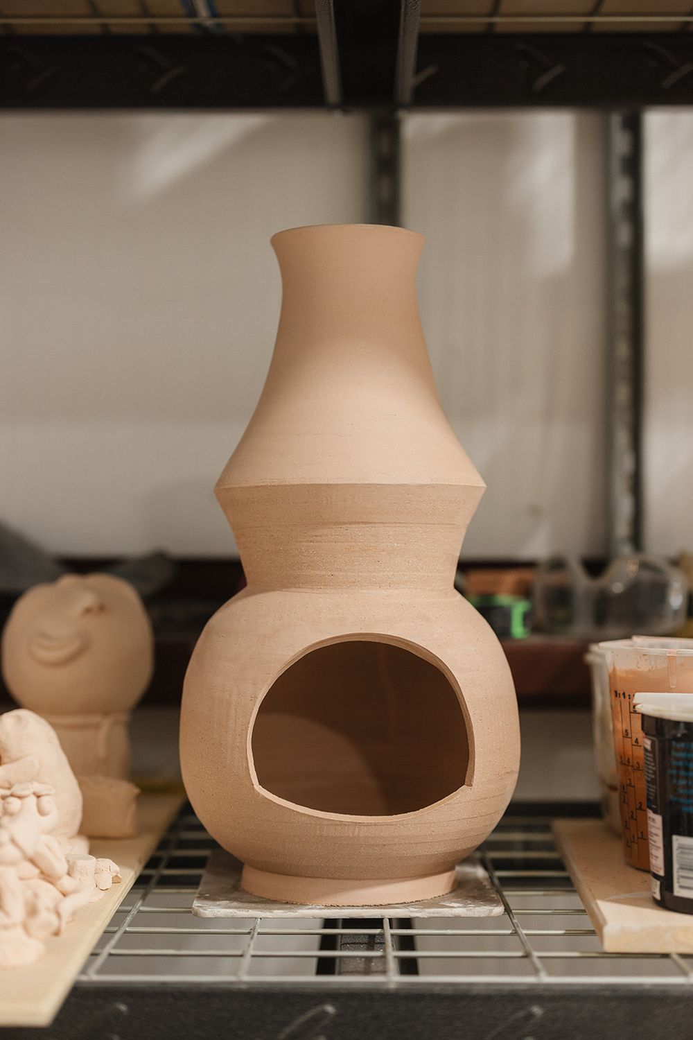 Unglazed clay pottery chiminea or lantern with rounded base and tall neck sits on metal shelf in workshop.