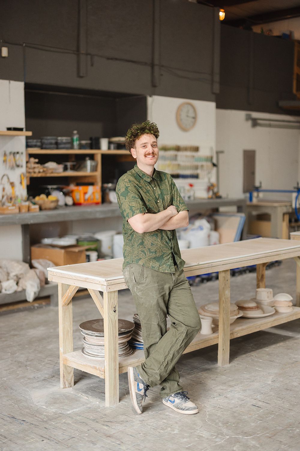 Artist in green workwear leans casually against wooden workbench in spacious pottery studio workshop.