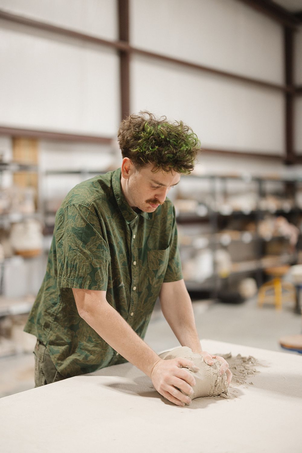 Person in green shirt works carefully with clay on a white table at a ceramics studio.