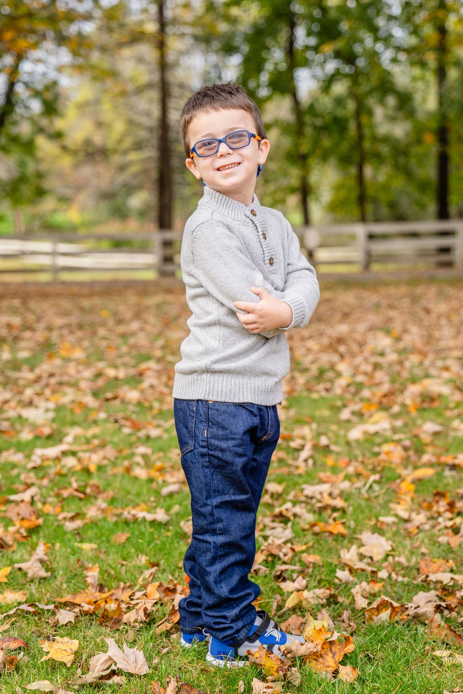 Kid wearing glasses and a gray sweater stands among fallen autumn leaves on grass with a wooden fence in the background. North Shore, MA